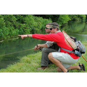 Two anglers kneeling on a grassy riverbank, one pointing across the water